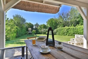 An outdoor dining area with a wooden table, a thatched roof, and a view of a garden with a wooden play structure and a lantern.