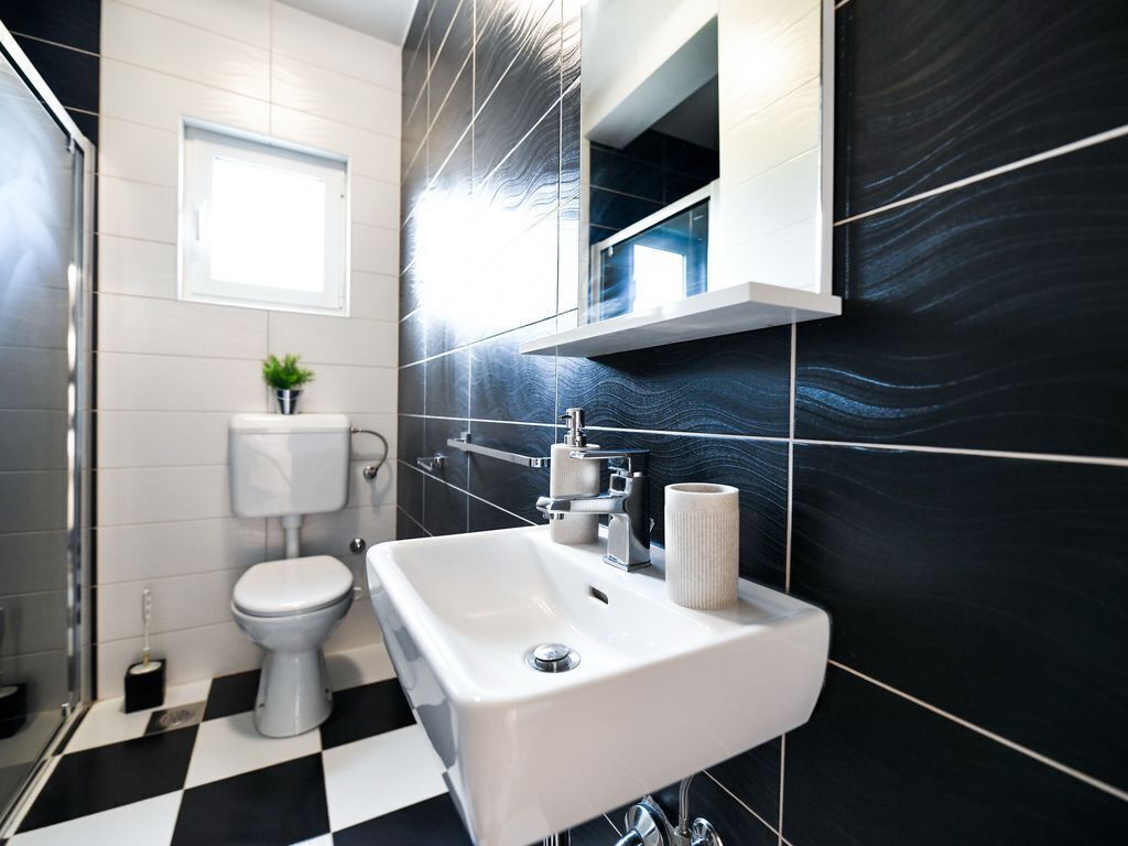 Ein modernes Badezimmer mit einem schwarz-weiß karierten Boden, einem weißen Waschbecken, einer Toilette und einem Fenster über der Toilette.