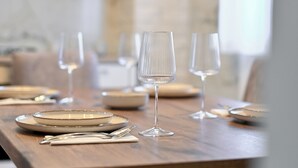Una mesa de comedor dispuesta con platos, cubiertos y copas de vino, con un muro de ladrillo y un armario blanco al fondo.