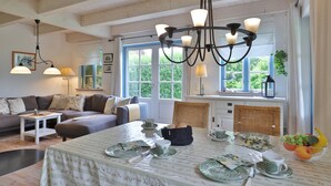 A cozy living room with a dining area, wooden beams on the ceiling, and a large window with a view of greenery.