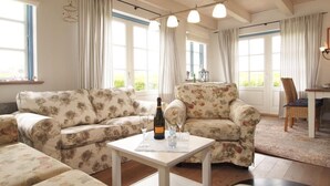 Een gezellige woonkamer met sofabekleding met bloemenprint, een klein tafeltje met een fles en glazen, en een eethoek op de achtergrond.