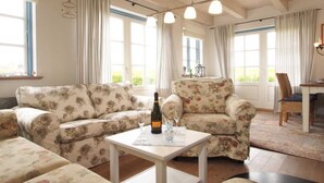 Een gezellige woonkamer met bloemenprintsofa's, een witte tafel met fles en glazen, en grote ramen met gordijnen.