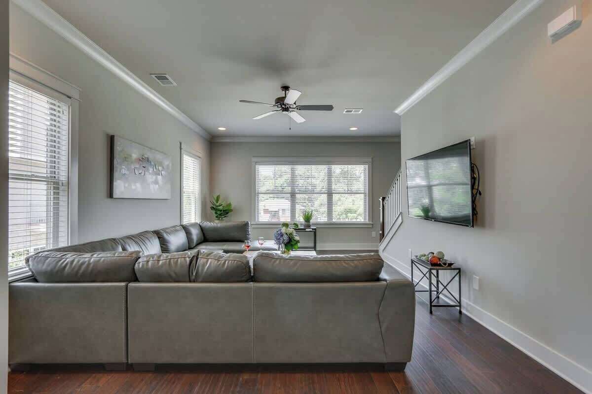 A modern living room with a sectional sofa, a ceiling fan, a flat-screen TV mounted on the wall, and a large window providing natural light.