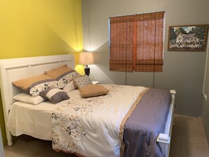 A bed with a floral bedspread and multiple pillows, a bedside lamp, and a window with bamboo blinds.
