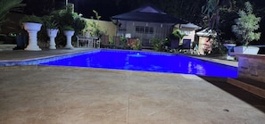 A swimming pool with a tiled deck area, surrounded by potted plants and a building in the background. - Cottage (St Joseph)