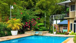 Une piscine bordée de pierres, entourée d’une végétation luxuriante, et un bâtiment à deux étages doté d’un balcon.