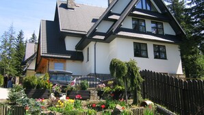 Une maison à deux étages, dotée d’un toit à forte pente, d’une cheminée bien visible et d’une clôture en bois qui entoure la propriété.