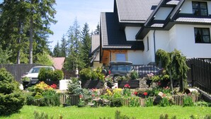 Une maison à deux étages avec un toit en pente, une voiture garée dans l’allée et un jardin où poussent différentes plantes et fleurs.