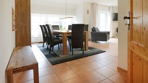 A dining area with a wooden table, chairs, and a rug. There is a wooden door on the right and a window with curtains in the background. - Sonnenblick holiday house (Kalkhorst)