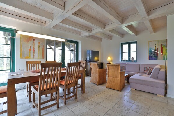 A spacious living area with a wooden ceiling, a dining table with chairs, a sofa, and a television.