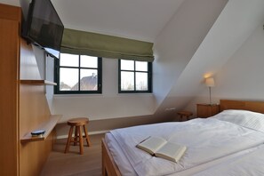 A bedroom with a bed, a TV, two windows, a lamp, and a stool. - Traveblick, Germany (Kalkhorst)