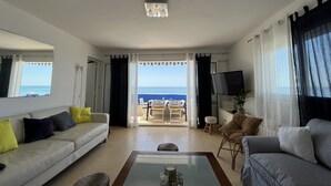 Een woonkamer met een bank, fauteuils, een salontafel en een televisie. Door de open gordijnen is een balkon met uitzicht op zee te zien.