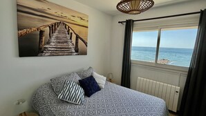 Een slaapkamer met een bedovertrek met patroon, uitzicht op zee door een raam, en een schilderij van een houten brug aan de muur.