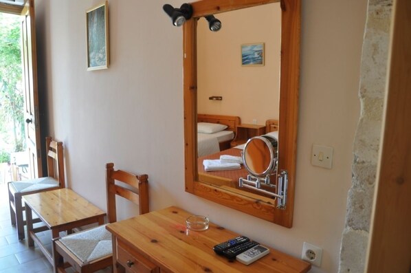 A room with a wooden desk, a chair, a bed with a wooden headboard, a large mirror, and a small table with a remote control and a glass.
