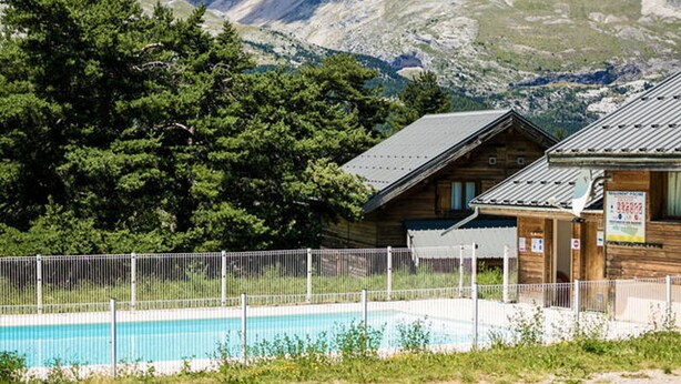 Una cabaña de madera con piscina, rodeada por una cerca, y montañas al fondo.