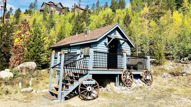 Una cabaña rústica de troncos con un porche y escalones, rodeada de árboles y un paisaje rocoso.