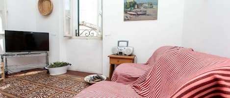 Una habitación con televisión, radio, alfombra estampada, cama con ropa de cama a rayas, ventana con vistas y un cuadro en la pared.