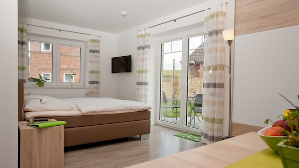 A modern bedroom with a bed, a TV on the wall, a window with curtains, a balcony with a table and chairs, and a green mat on the floor. - Apartment Alma - Apartment Lühe (Jork)