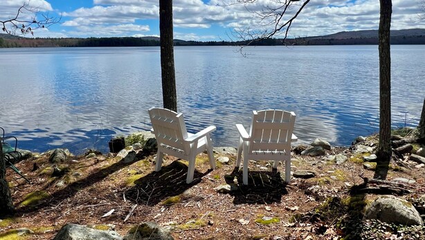 Zwei Adirondack-Stühle mit Blick auf einen stillen See bei blauem Himmel.