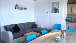 Moderni olohuone, jossa on harmaa sohva, siniset tuolit, puinen pöytä ja avoimen oven kautta näkyvä keittiötila.