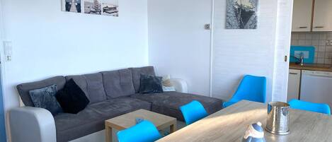 A modern living room with a grey sofa, blue chairs, a wooden table, and a kitchen area visible through an open door.