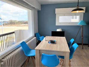 Une pièce avec une table en bois et des chaises bleues, une fenêtre donnant sur la plage, un radiateur et un lampadaire.