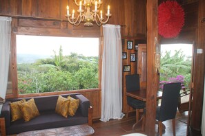 A wooden room with a sofa, dining table, and chairs. A large window offers a view of greenery outside.
