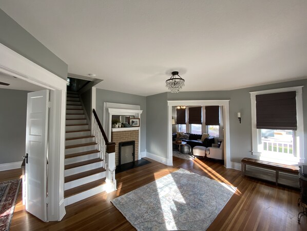 A spacious living area with a staircase, a fireplace, and a large window with blinds.