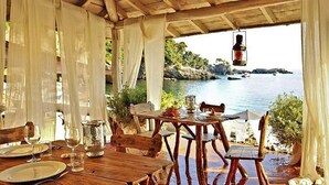 Un espace de restauration en plein air avec des tables et des chaises en bois, des rideaux blancs et une vue sur la mer.