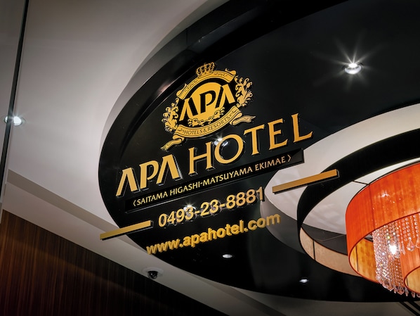 A sign for APA HOTEL with a crown logo, contact number, and website URL.