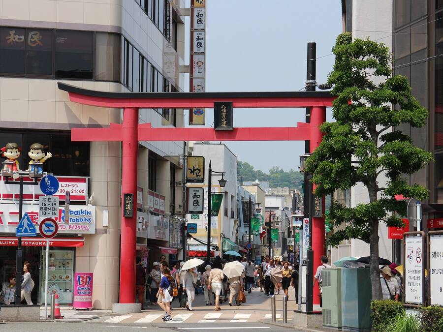 A busy urban street with a red torii gate, storefronts, and pedestrians.