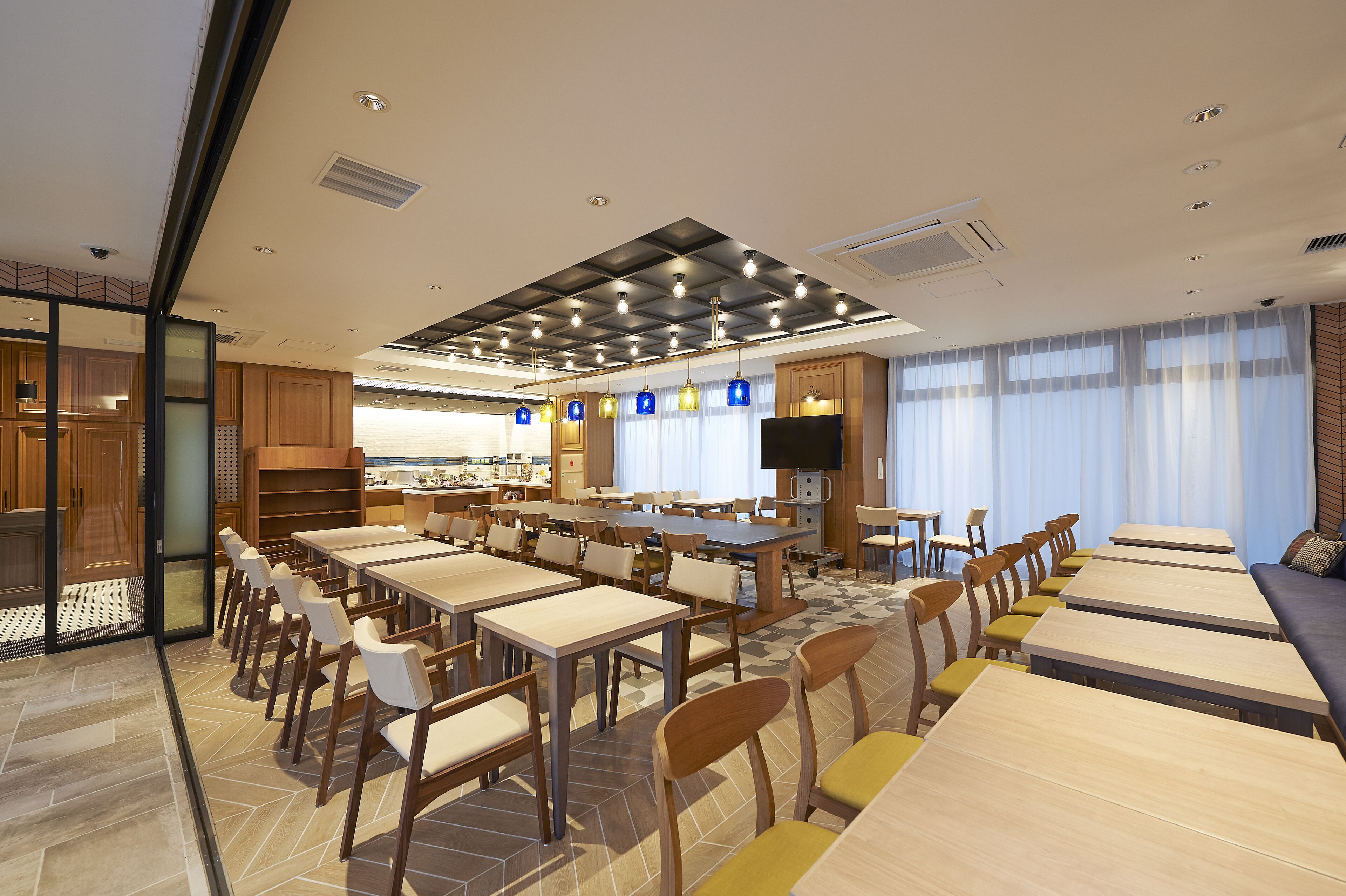 A spacious dining area with multiple tables and chairs, a kitchen in the background, and a large window with curtains.