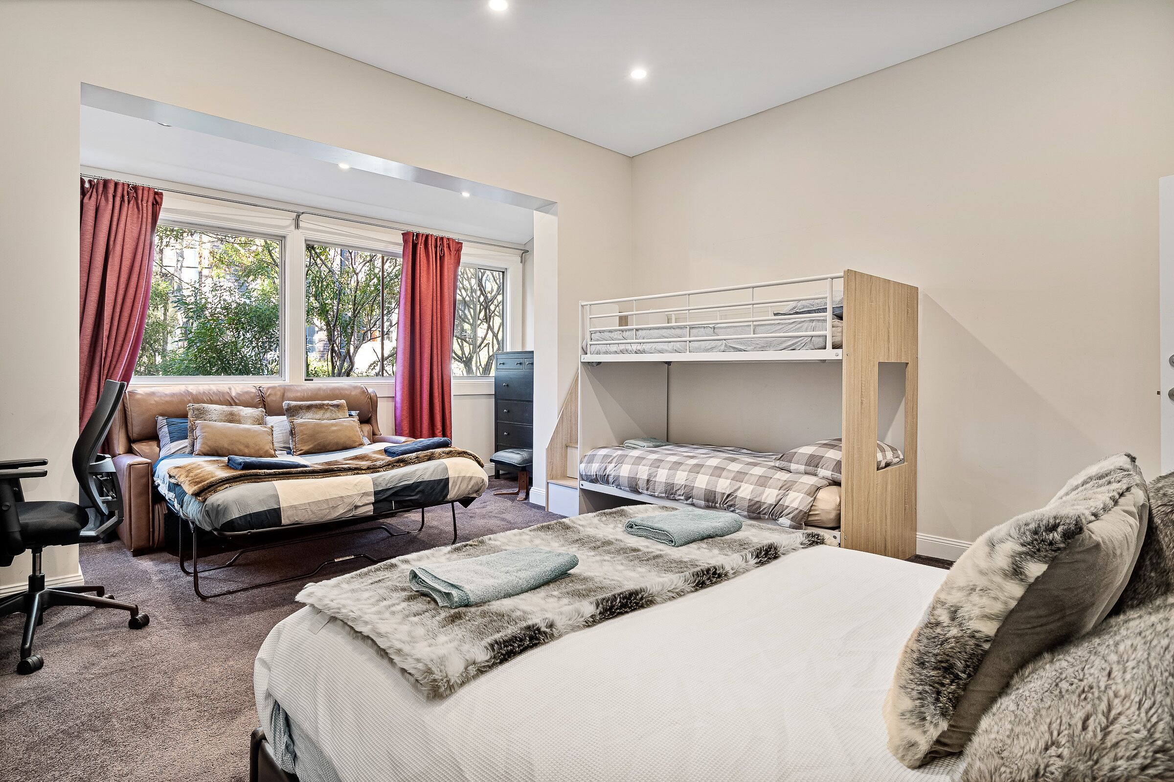A bedroom with a large bed, a smaller bed, a desk, and a chair. There is a window with curtains and a view of trees.