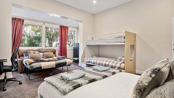 A bedroom with a large bed, a smaller bed, a desk, and a chair. There is a window with curtains and a view of trees.