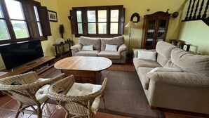 Un salon comprenant un téléviseur, deux canapés, une table basse ronde, des fauteuils en osier, une lampe et une fenêtre avec des volets.