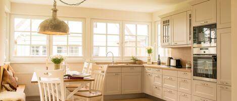 A kitchen with white cabinets, a wooden dining table, and chairs. There is a window with a view of the outside.