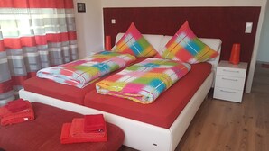 Een net opgemaakt bed met kleurrijke kussens en een rode deken, een klein nachtkastje met een lamp, en een houten vloer.