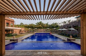 Una piscina con una profundidad de 38 centímetros, rodeada de pérgolas de madera, mobiliario de exterior y un edificio de piedra al fondo.