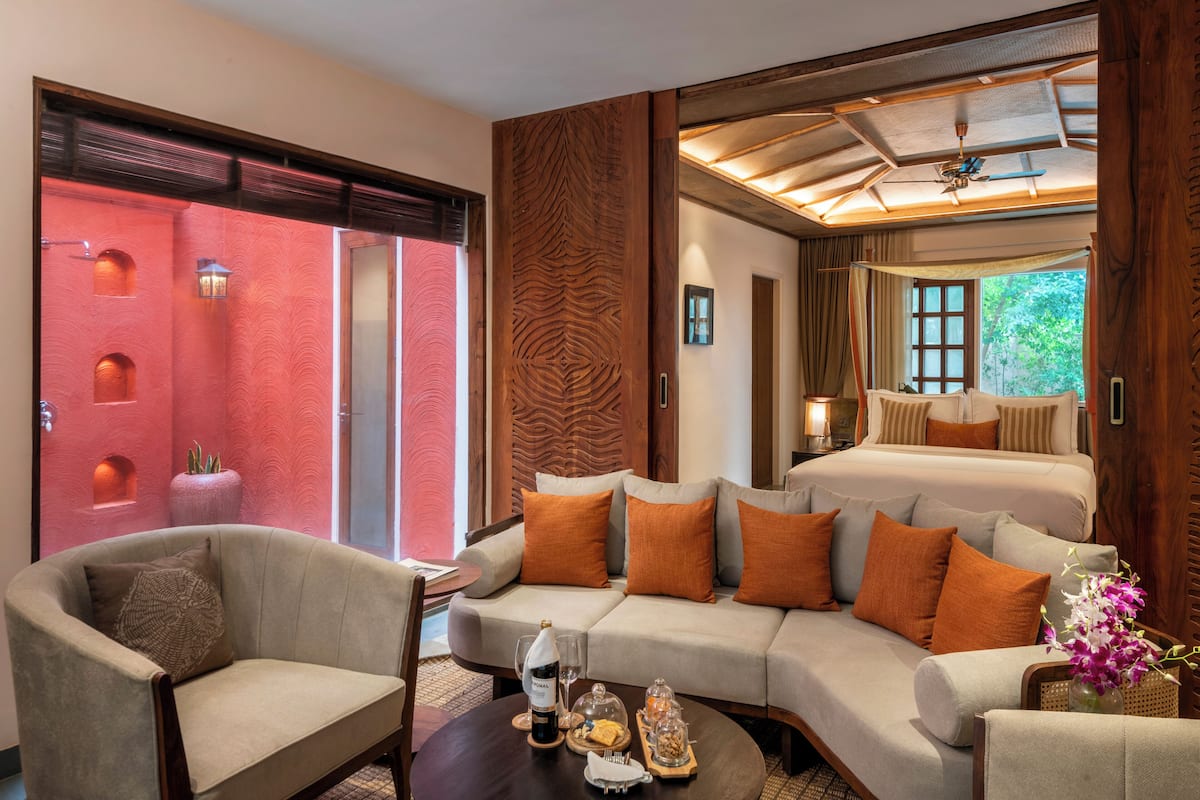 A modern hotel room with a large sofa, a dining table with a bottle and glasses, and a bed with orange pillows.