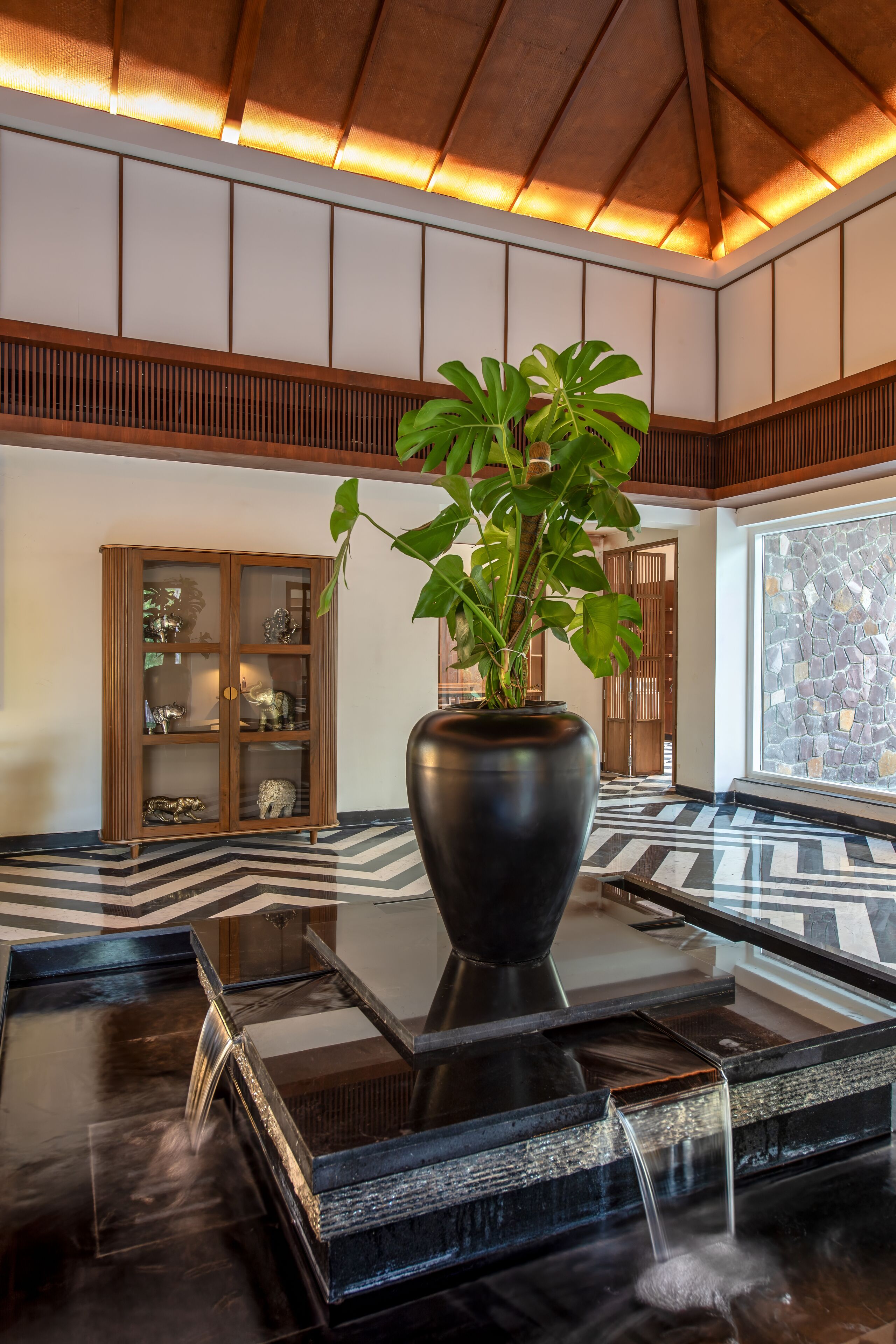 Un hall d’entrée avec un sol en marbre noir, un grand vase noir contenant une plante, un meuble en bois doté de portes en verre et un mur en pierre.