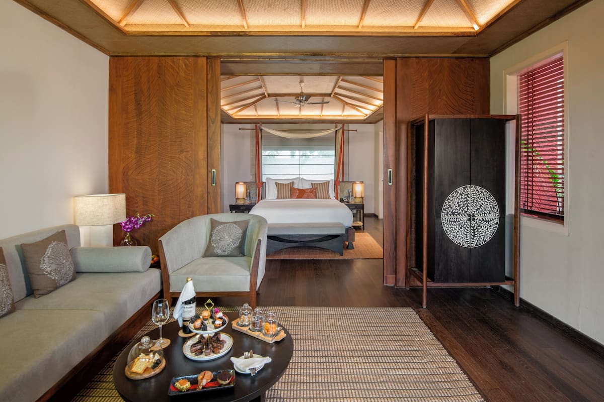 A modern hotel room with a bed, a sofa, a dining table with food, and a decorative cabinet.