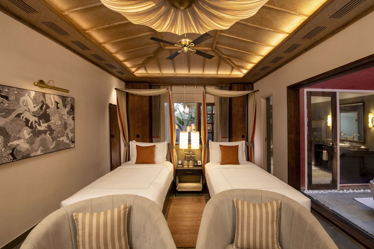 A hotel room with two beds, a ceiling fan, a painting on the wall, and a view of the outside.