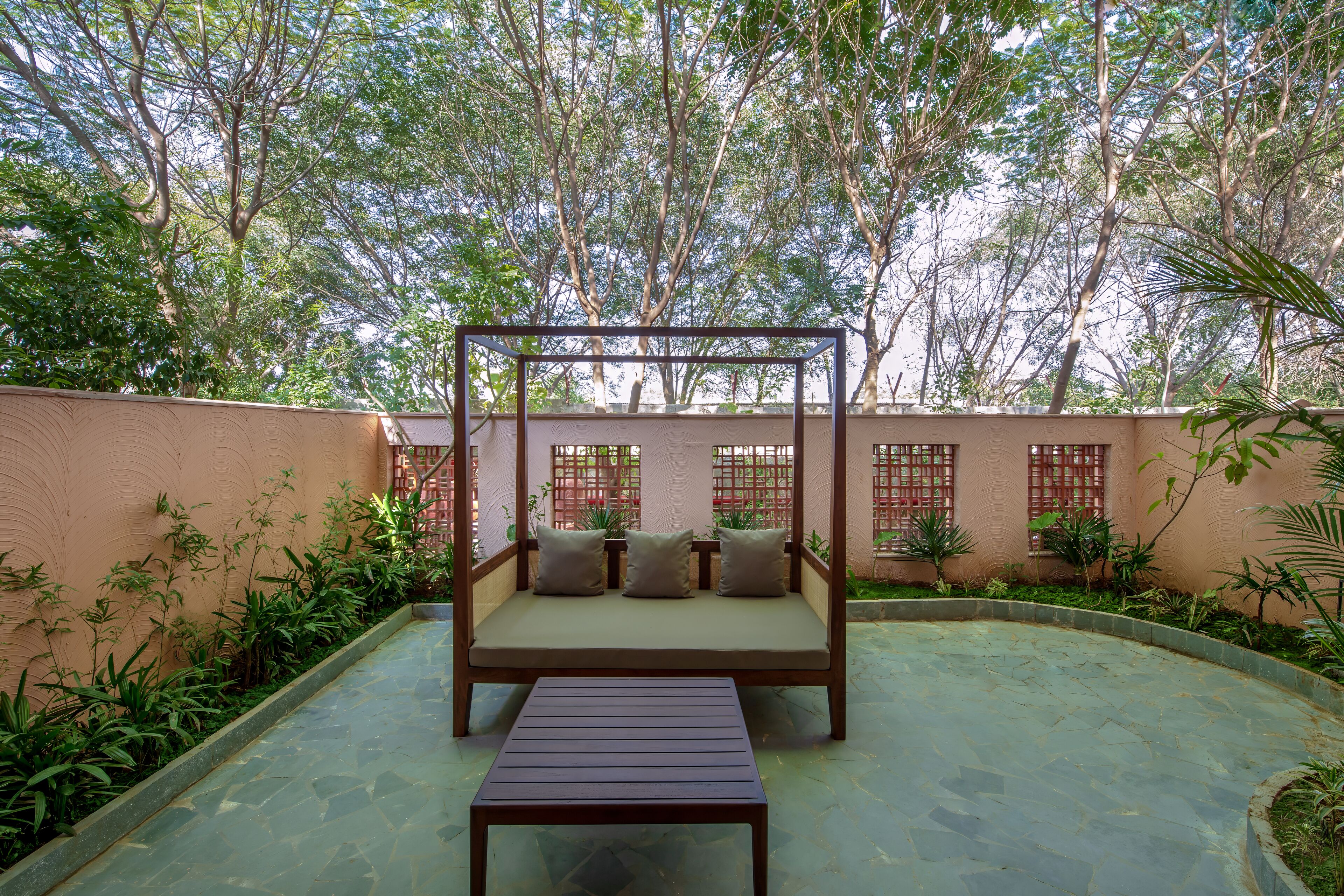 A garden patio with a wooden bench and a low table, surrounded by lush greenery and trees.