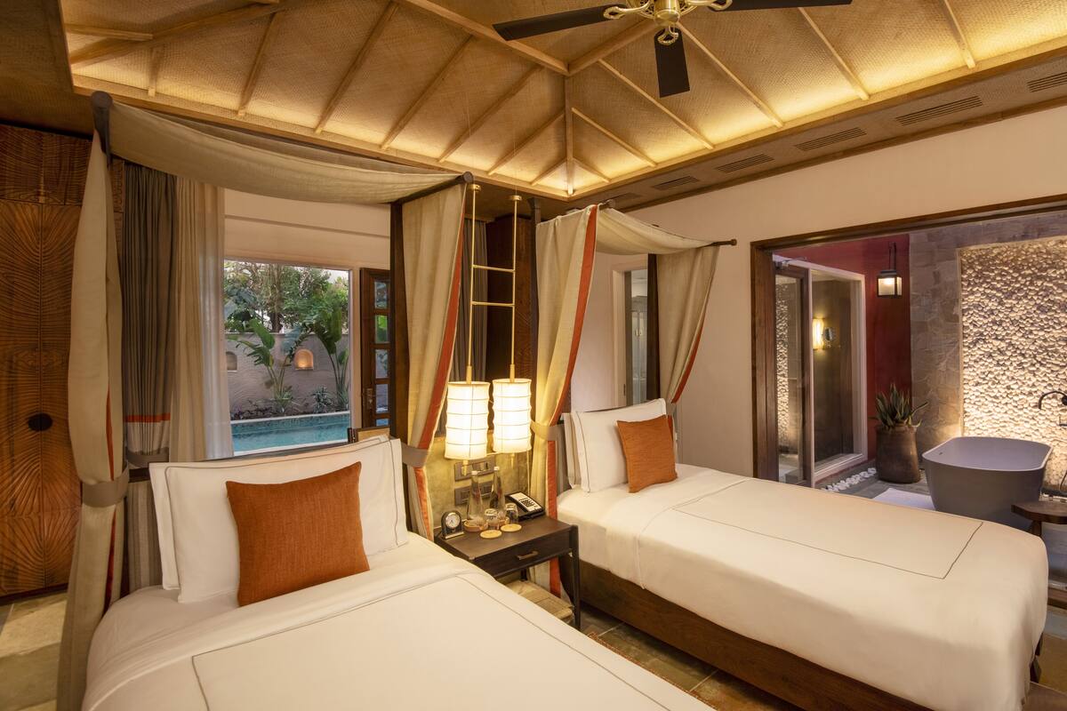 A hotel room with two beds, a nightstand with a lamp, a window with curtains, and a view of the outdoors.