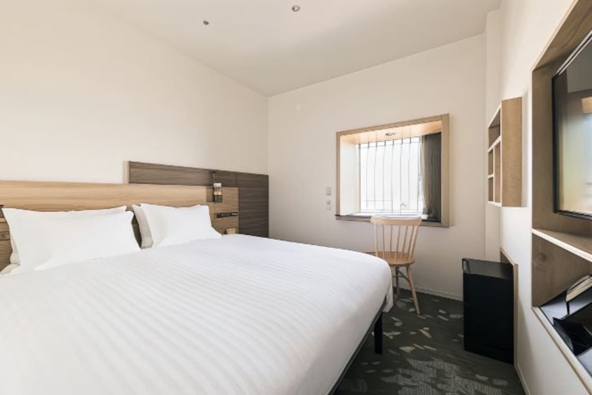 Habitación de hotel con una cama grande, cabecero de madera, un escritorio pequeño, una silla, un televisor y una ventana con persianas.