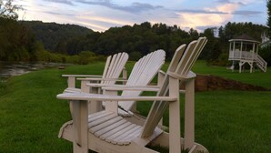 Dos sillas Adirondack en una terraza de madera con vista a un estanque y un pabellón al fondo.