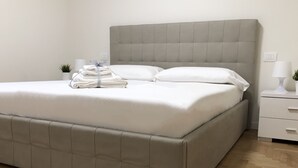Een bed met een gestoffeerd hoofdbord, een nachtkastje met een lamp en een kleine plant.