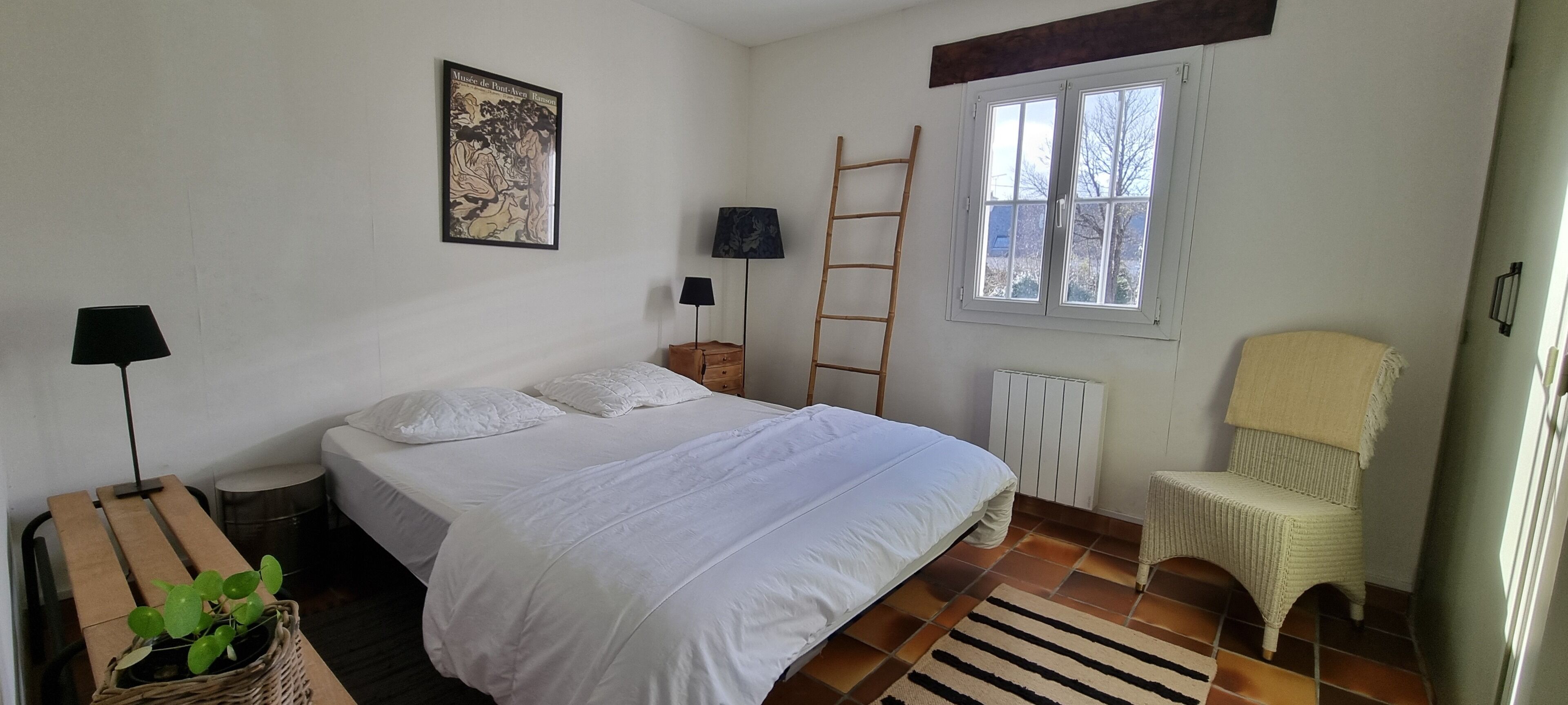 Un dormitorio con cama, ventana, escalera, silla, radiador y lámpara.