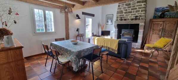 Un salon chaleureux avec une cheminée en pierre, une table à manger avec des chaises et un canapé recouvert d’un plaid jaune.