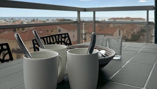 A balcony with a table set for two, featuring white mugs and a glass of water, overlooking a cityscape. - Very nice apartment with sea view and private parking 4SUF502 (LE BARCARES)